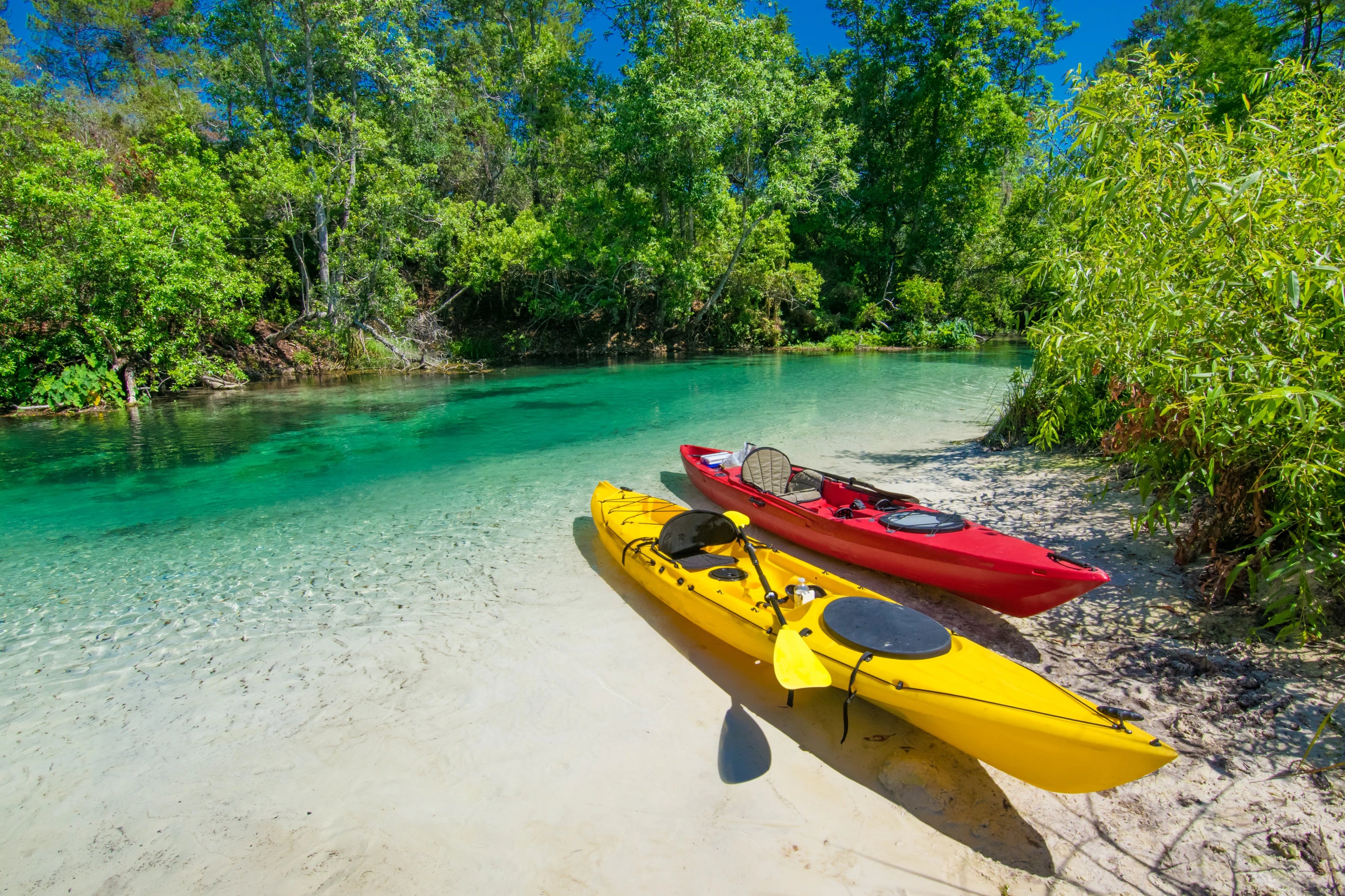 Two kayaks, one yellow and one red, on a sandy beach with clear turquoise water and greenery.